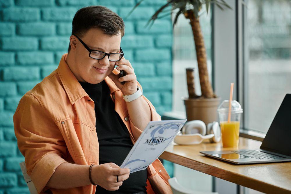 Person sitting at café table, holding menu while on phone. Laptop and juice on table.